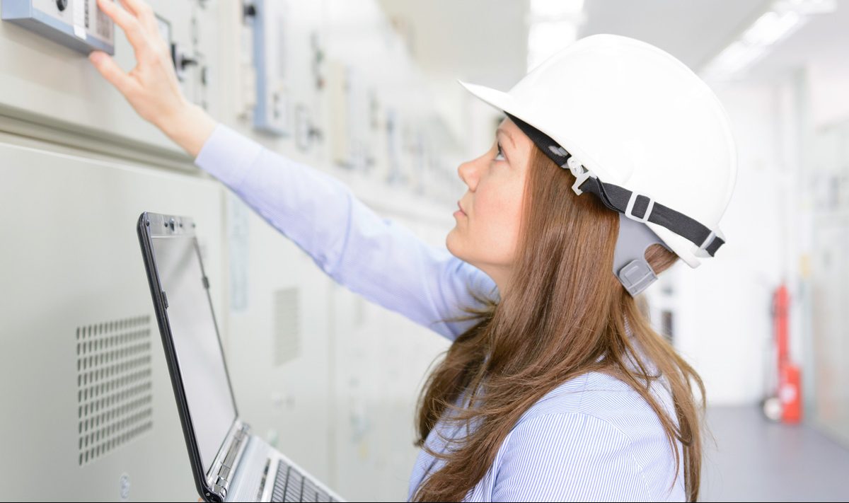 Female engineer checking equipment in power substation wearing white safety hat and holding laptop in left hand.