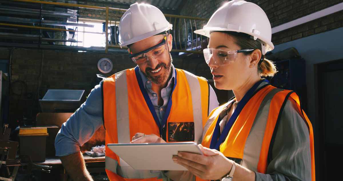 One male and one female engineer using a digital tablet together in an industrial place of work. They are both wearing safety hats and high visibility clothing.