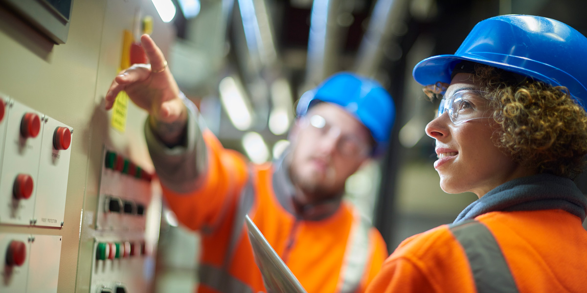 Two industrial service engineers conduct a safety check of a control panel and boiler room at a power station. They are both wearing safety equipment and are looking at a control panel.