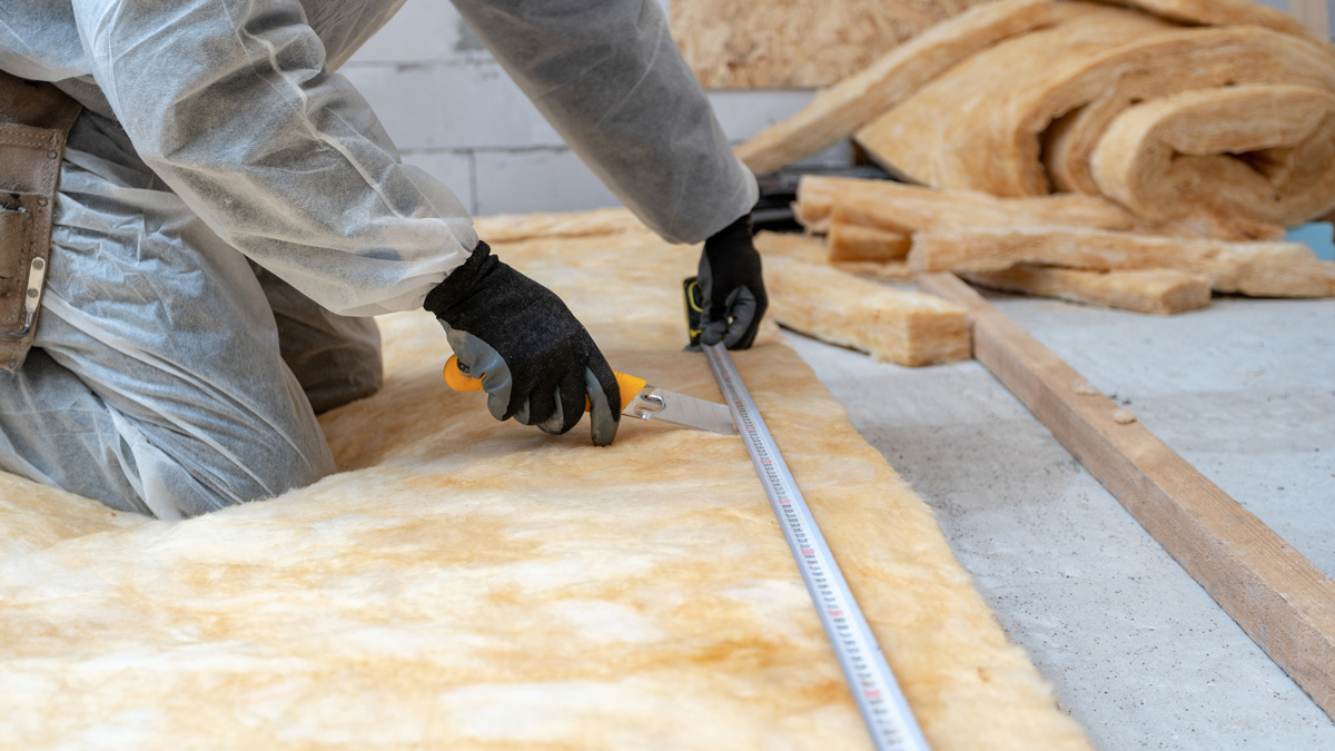Cropped view of professional workman in protective workwear using knife and holding measuring tape over material, installing thermal insulation rock wool under the roof.
