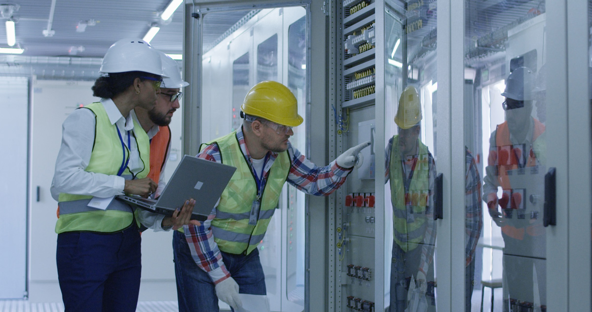 Group of multiethnic men and woman in hardhats working in hall of solar plant control centre having discussion between racks.